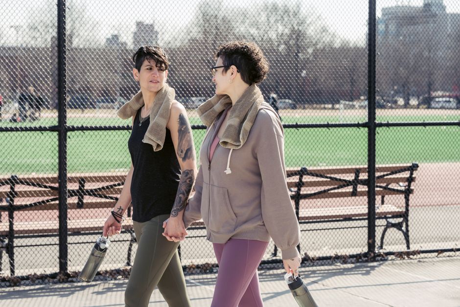 Two women walking hand in hand on a sunny outdoor sports court, behind a chain-link fence, with a green sports field and benches in the background. The woman on the left has short dark hair, casual bl