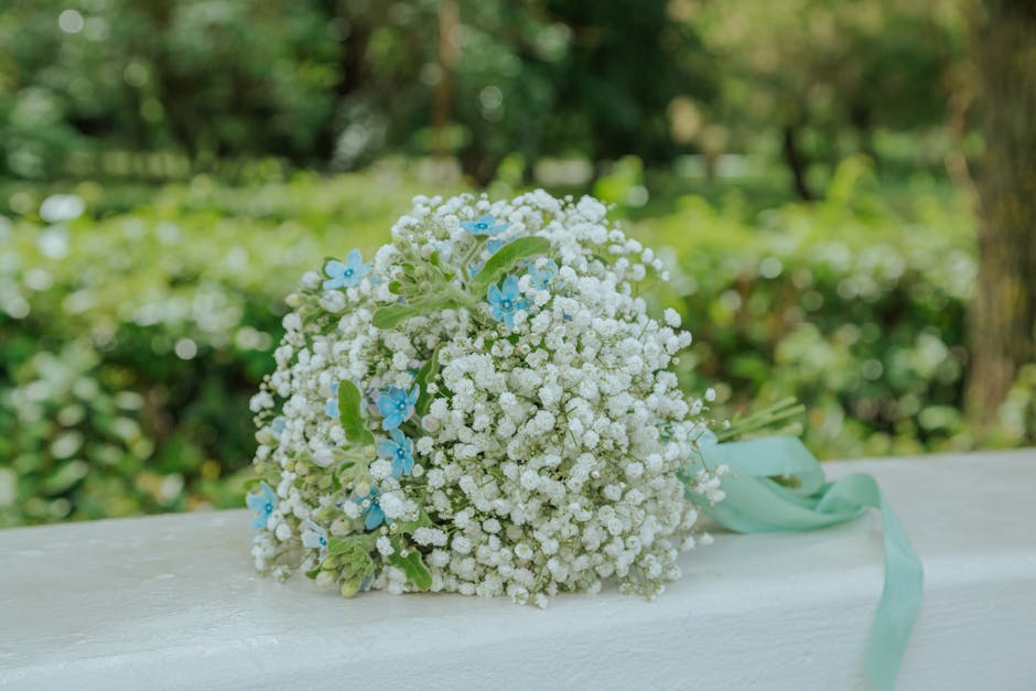 A bouquet featuring white gypsophila (baby's breath) with small light blue flowers interspersed, arranged in a rounded, lush style. The bouquet is tied with a soft mint green ribbon that drapes over a