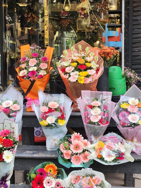 A vibrant display of freshly arranged flower bouquets outside a floral shop. The bouquets feature a variety of blooms including pink gerbera daisies, white chrysanthemums, yellow roses, and pink carna