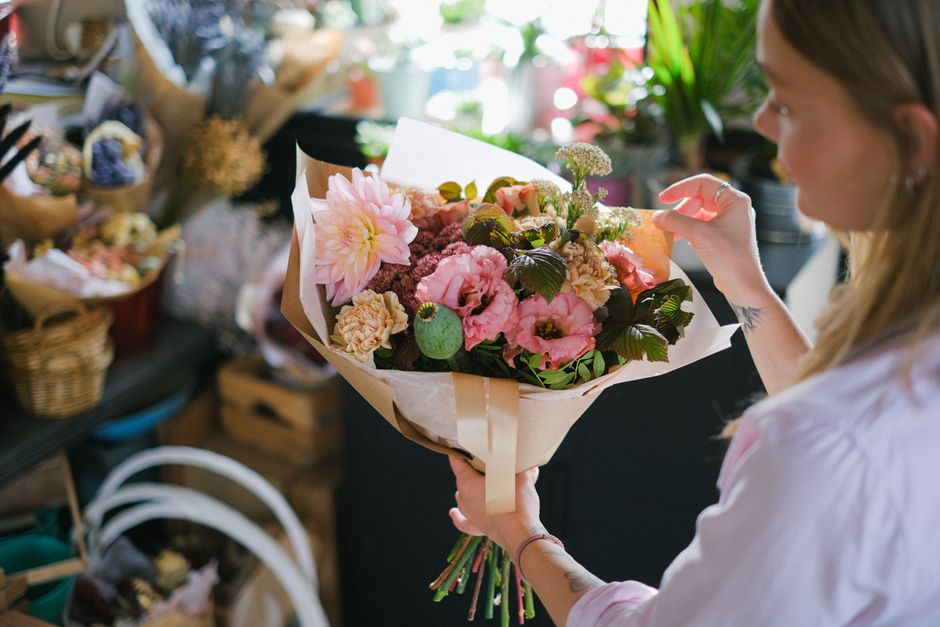 A woman with light brown hair and a light pink shirt is holding a large bouquet of fresh flowers, including pink dahlias, roses, and lisianthus, wrapped in beige and white paper with a satin ribbon. S